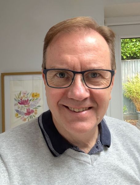 A man with short light brown hair and glasses is smiling at the camera. He is wearing a grey jumper over a collared shirt, standing indoors with a floral painting and a window with greenery behind him.
