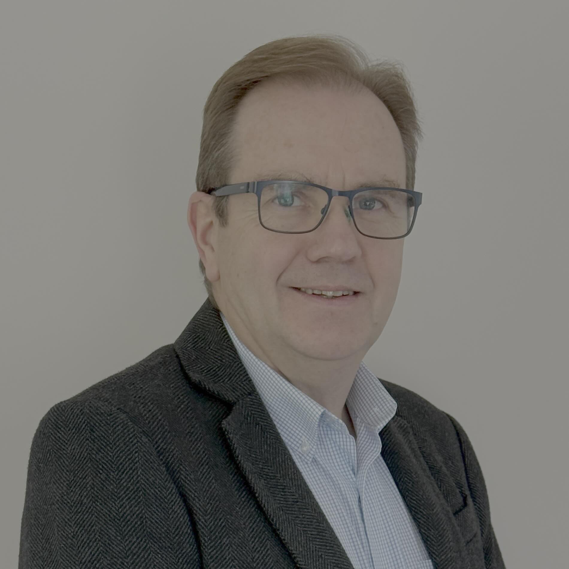 A middle-aged man with short light brown hair and glasses, wearing a dark blazer over a light blue shirt, smiling and posing in front of a plain white background. A middle-aged man with short light brown hair and glasses, wearing a dark blazer over a light blue shirt, smiling and posing in front of a plain white background.